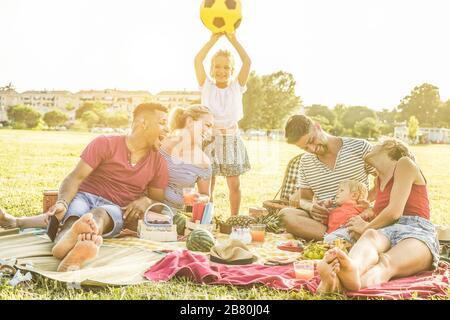 Les familles heureuses qui pique-niquent dans le parc de la ville - les jeunes amis s'amusent avec leurs enfants en été en mangeant, en buvant et en riant ensemble - Love A. Banque D'Images