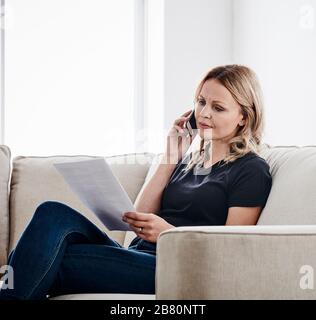 Photo d'une femme assise sur un canapé tenant un document et parlant sur un téléphone portable Banque D'Images