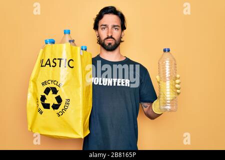 Homme volontaire hispanique portant un sac de transport de t-shirt volontaire avec des bouteilles en plastique avec une expression sûre sur la pensée intelligente du visage serio Banque D'Images