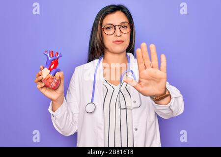 Jeune médecin spécialiste de cardiologie femme tenant le coeur médical sur le fond de pruple avec main ouverte faisant signe d'arrêt avec sérieux et confiant expres Banque D'Images