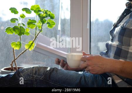 Un homme est assis sur un seuil de fenêtre avec une tasse de café et un livre pendant la quarantaine Banque D'Images