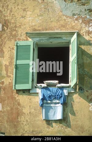 Le lavage s'est accroché à sécher à partir d'une fenêtre dans un bâtiment à Parga, Preveza, Epirus, Grèce. Il est maintenu en place par des coussins. Les volets verts jettent une ombre sur le mur inégal et ocre de la maison. Banque D'Images