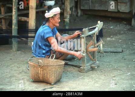 Lao femme, assis sur le sol, filant sur sa maison fait, main a lancé, roue de rotation en bois à l'extérieur de sa maison. Elle porte une robe traditionnelle et possède un panier en osier sur son côté. Les longueurs de fil noir fini s'étirent derrière elle. Banque D'Images