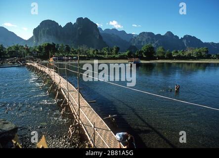 Pont en bois et natte de paille tissée sur la rivière Nam Song à Vang Vien (Vang Vieng), dans la province de Vientiane, au Laos. Le toit de chaume et les parasols multicolores d'un café se trouvent sur la rive éloignée. Les enfants jouent dans l'eau et un camion est lavé. Il y a des montagnes couvertes d'arbres, des montagnes karstiques et un ciel bleu clair derrière. Banque D'Images