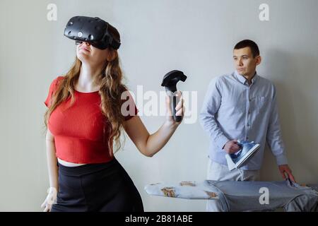 Une fille dans des lunettes de réalité virtuelle est en train de jouer. Son mari à cette fois strober le linge de maison. Banque D'Images