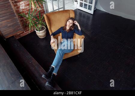 Une fille dans une chemise et un Jean se trouve sur un fauteuil jaune. Le sol de la chambre est en bois. À proximité se trouve une fleur intérieure. Banque D'Images