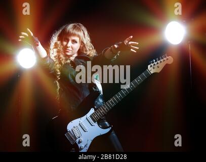 Guitariste fille avec guitare noire et blanche, dirige ses mains vers la caméra. Derrière deux projecteurs. L'arrière-plan est noir Banque D'Images