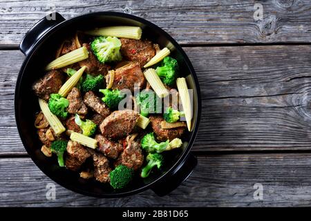 Morceaux de porc rôtis dans le four mélangés avec du brocoli et des épis de maïs sur un plat noir sur un vieux fond en bois, vue de dessus, espace de copie Banque D'Images