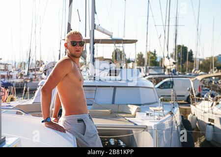 Un jeune homme heureux se sent heureux sur la marina près du catamaran de bateau à voile de luxe pendant les vacances d'été. Modèle masculin caucasien. Banque D'Images