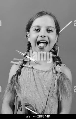 Girl détient sac d'école dans les mains et en forme d'coeur cookie sur la langue. Fille de l'école avec un visage heureux isolé sur fond saumon. Concept Retour à l'école. Kid en robe rose avec des tresses et crayons de couleur. Banque D'Images