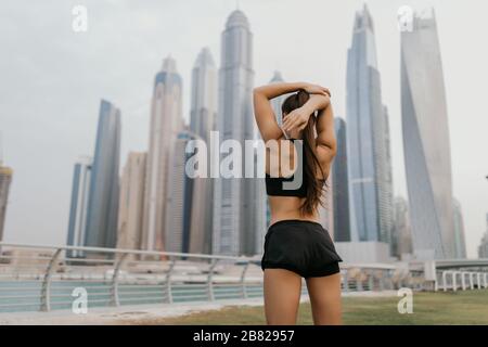 Jeune fille sportive portant des vêtements de sport roses qui s'étirent à proximité des gratte-ciel, se préparant à l'entraînement, à l'entraînement en extérieur, se réchauffant après ou avant la fi Banque D'Images