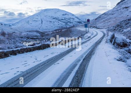 Traces de pneus et empreintes de pas sur route enneigée courbant à travers les montagnes à Glencoe, Highlands, Écosse en hiver Banque D'Images