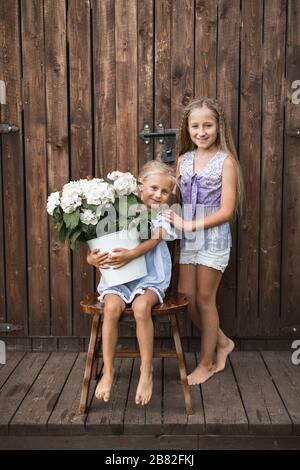 Jolie petite fille blonde souriante avec grand bouquet de fleurs d'hydracea, assise sur la chaise contre le fond de la grange en bois, tandis que sa sœur plus ancienne Banque D'Images