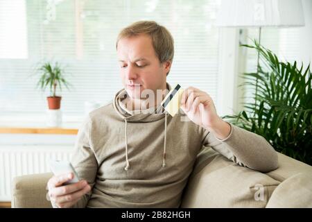 Un homme adulte heureux assis à la maison sur un canapé avec ordinateur portable et carte de crédit. Payer des factures et des commandes. Concept de commerce électronique et d'achat en ligne. Banque D'Images