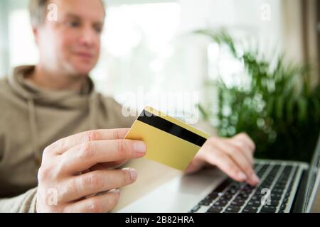 Un homme adulte heureux assis à la maison sur un canapé avec ordinateur portable et carte de crédit. Payer des factures et des commandes. Concept de commerce électronique et d'achat en ligne. Banque D'Images