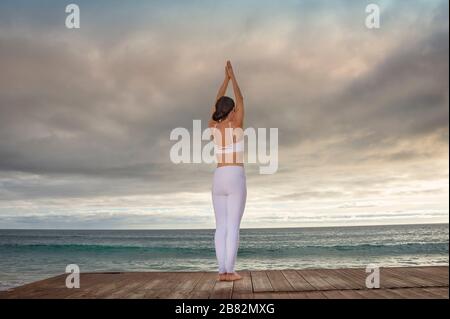 femme debout près de l'océan pratiquant le yoga, vue arrière, ciel dramatique. Banque D'Images