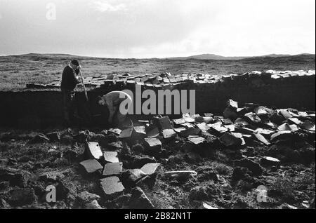 Deux hommes qui coupent de la tourbe sur une étendue de terre au Loch Porlan, sur l'île de l'Uist du Nord, dans les Hébrides extérieures, en Écosse. La coupe de la tourbe était une méthode traditionnelle de collecte de carburant pour l'hiver dans les régions peu peuplées de la côte ouest de l'Écosse et des îles. La tourbe a été séchée et utilisée dans les feux et les fours. Banque D'Images