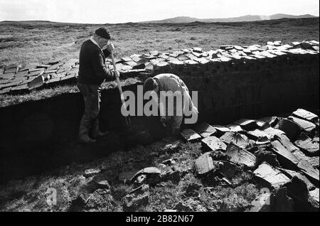 Deux hommes qui coupent de la tourbe sur une étendue de terre au Loch Porlan, sur l'île de l'Uist du Nord, dans les Hébrides extérieures, en Écosse. La coupe de la tourbe était une méthode traditionnelle de collecte de carburant pour l'hiver dans les régions peu peuplées de la côte ouest de l'Écosse et des îles. La tourbe a été séchée et utilisée dans les feux et les fours. Banque D'Images