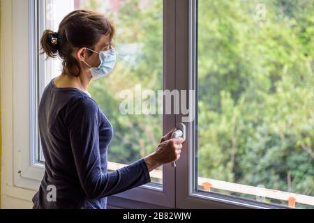 Une femme mise en quarantaine à la maison, portant un masque médical et une tenue décontractée, est en veille devant une fenêtre fermée, en regardant dans l'espace. Banque D'Images