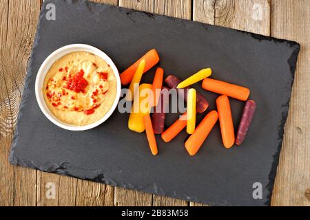 Faites tremper les carottes arc-en-ciel avec de l'houmous sur un serveur d'ardoise sur un fond rustique en bois Banque D'Images