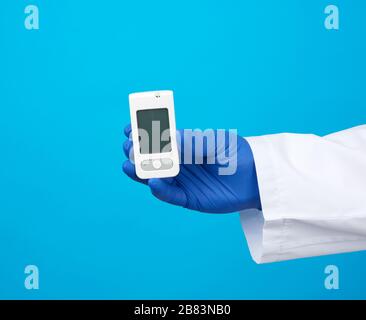 la main masculine avec des gants stériles bleus contient un glucomètre de sang blanc pour mesurer la glycémie, fond bleu Banque D'Images