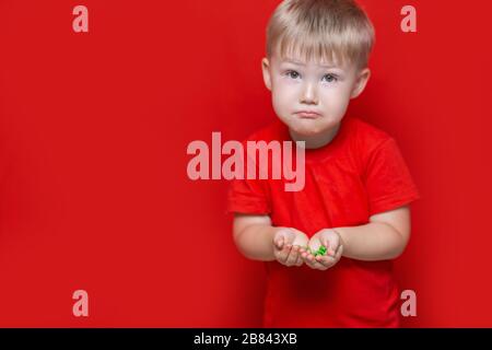 petit garçon triste enfant tenir beaucoup de pilules dans les mains. t-shirt rouge, fond rouge dangereux concept, comprimés et bébé Banque D'Images