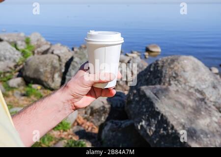 Tasse de café en papier du matin avec café chaud savoureux chez les hommes Main à l'extérieur sur fond de mer.main tient un papier de carton blanc tasse pour boissons à emporter Banque D'Images