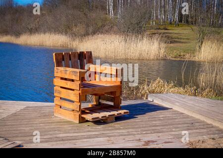 Chaise rustique faite à la main à partir de palettes en bois sur terrasse en bois donnant sur une rivière ou un lac avec des roseaux au soleil de printemps Banque D'Images