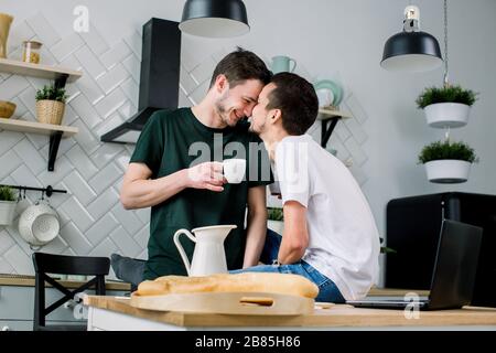 Un couple gay heureux prenant le petit déjeuner dans la cuisine et utilisant un ordinateur portable. Les hommes homosexuels pleins de plaisir se embrassent, font face à face tout en passant du temps à la maison. Banque D'Images