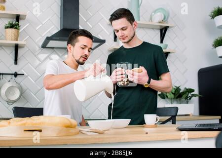 Un couple gay heureux prépare le petit déjeuner dans la cuisine le matin. Beau homme mettant des céréales de petit déjeuner dans des bols blancs, tandis que son partenaire verser du lait dans Banque D'Images