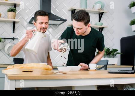 Un couple gay heureux prépare le petit déjeuner dans la cuisine le matin. Beau homme mettant des céréales de petit déjeuner dans des bols blancs, tandis que son partenaire verser du lait dans Banque D'Images