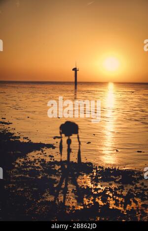 Coucher de soleil doré dynamique derrière des nuages bas suspendus au-dessus de la mer du nord sur la côte néerlandaise. Un appareil photo hors de la mise au point sur un trépied attend la photo Banque D'Images