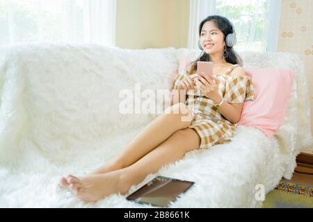 Les femmes asiatiques ont l'air mignonnes portant un casque et écoutant de la musique connectée au smartphone. Dans sa chambre. Assis sur le canapé avec une lumière blanche s Banque D'Images