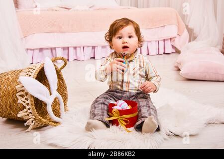 Rire bébé garçon avec des oreilles de lapin chassant pour des œufs de Pâques à la maison. Mignon petit garçon à poil dur assis sur le sol avec une boîte d'œufs de Pâques Banque D'Images