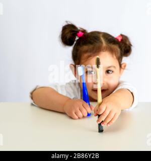 Petite belle fille tenant éco naturel de la brosse à dents en bambou et dans la brosse à dents en plastique bleu d'autre main. Zéro gaspillage, concept de style de vie durable. Focalisation sélective sur la brosse à dents en bambou. Banque D'Images