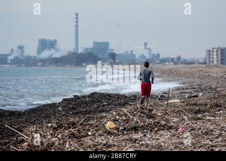 Une personne est en train de courir sur une plage pleine de ordures avec une usine polluante en arrière-plan Banque D'Images