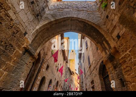 Une arche au-dessus de la rue via S. Matteo avec des drapeaux rouges sur les murs dans le centre historique de San Gimignano, province de Sienne, Italie, au coucher du soleil. Banque D'Images