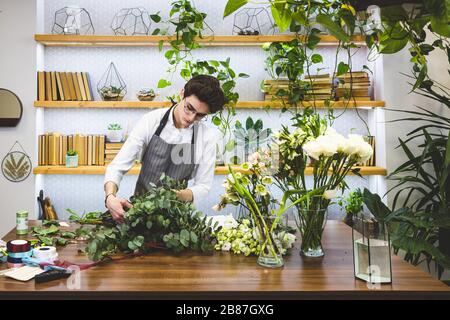 Beau jeune fleuriste masculin avec lunettes et tablier travaille dans un magasin de fleurs. Banque D'Images