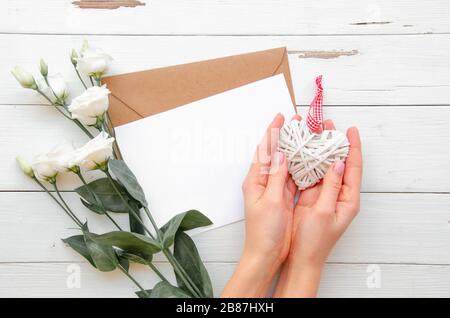 Mains féminines tenant le coeur en bois blanc fait main au-dessus d'un papier pour note, une lettre, et de belles fleurs de roses sur une table de bureau ligneuse blanche, vue sur le dessus, Banque D'Images