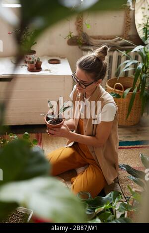 Jeune jolie femme dans des lunettes assis et transplanter de jeunes fleurs et plantes dans les pots dans la chambre Banque D'Images