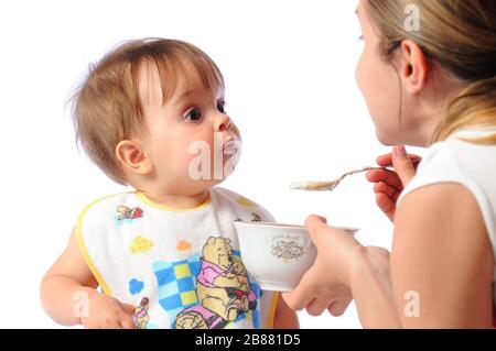 La mère nourrit la petite fille surprise de la nourriture de bébé de la cuillère. Soins parentaux et bonne enfance. Isolé sur fond blanc Banque D'Images