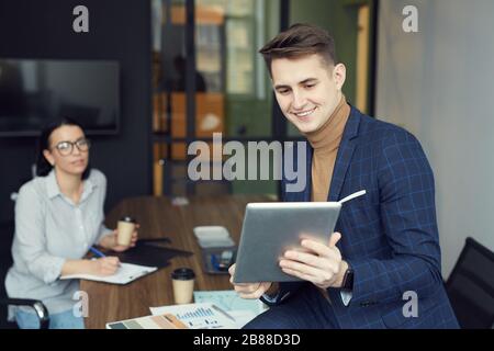 Jeune homme d'affaires tenant une tablette pc et l'utilisant pour la communication en ligne avec la femme d'affaires assis dans l'arrière-plan au bureau Banque D'Images
