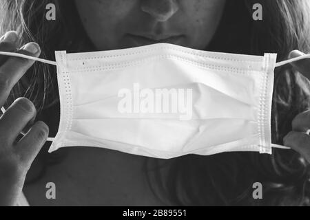 Photo en noir et blanc d'une jeune femme non reconnaissable mettant sur un masque médical blanc. Fond flou, mise au point sélective. Épidémie de coronavirus, quarantaine COVID-19. Concept médecin/infirmière. Banque D'Images