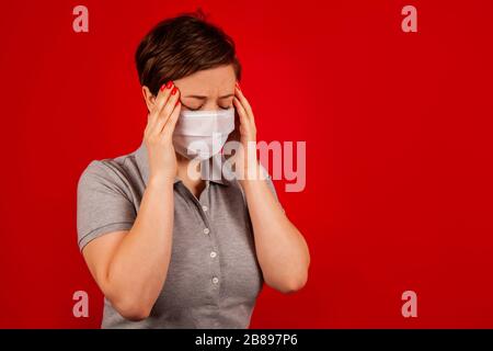 Portrait d'une femme dans un masque présentant un symptôme d'une maladie virale. La femme a mal à la tête. La femme dans un masque tient les mains près du temple tout en se tenant sur un fond rouge Banque D'Images