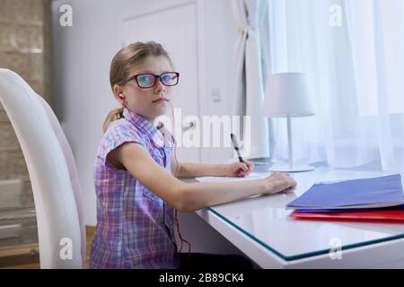 Fille enfant de 9 ans, 10 ans assis à la maison au bureau près de la fenêtre et écrire dans un ordinateur portable, écouteurs dans les oreilles, cours audio, technologie dans l'éducation Banque D'Images