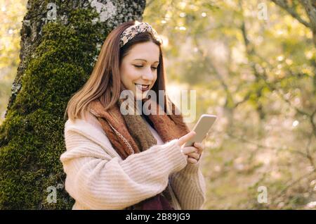 Jeune femme souriante, se penchant contre un arbre, utilisant son téléphone portable Banque D'Images