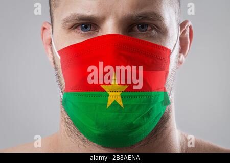 Jeune homme aux yeux douloureux dans un masque médical peint dans les couleurs du drapeau national du Burkina Faso . Protection médicale contre les maladies aéroportées, Banque D'Images