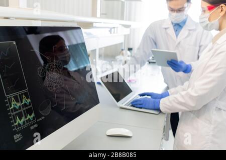 Écran d'ordinateur avec modèle de molécule en arrière-plan de deux travailleurs de laboratoire en blanchons, masques et gants étudiant l'information en ligne Banque D'Images