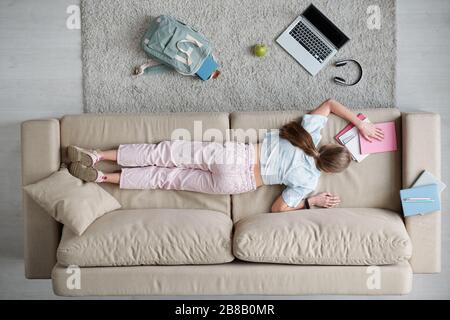 Vue d'ensemble de l'arrière d'une adolescente posée sur un canapé entouré de sac à dos, d'un ordinateur portable, d'un casque et de copybooks pendant les devoirs Banque D'Images