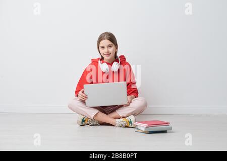 Jolie adolescente en tenue de casual traversant ses jambes tout en étant assise au mur et surfer dans le filet tout en étudiant à distance en quarantaine Banque D'Images